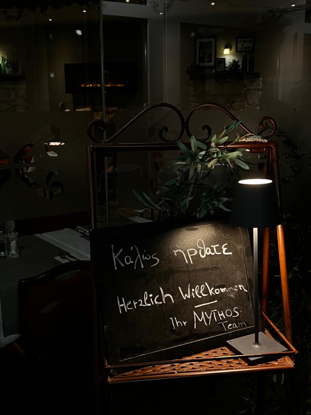 Eine schwarze Tafel mit weißer Schrift begrüßt freundlich auf Deutsch und Griechisch